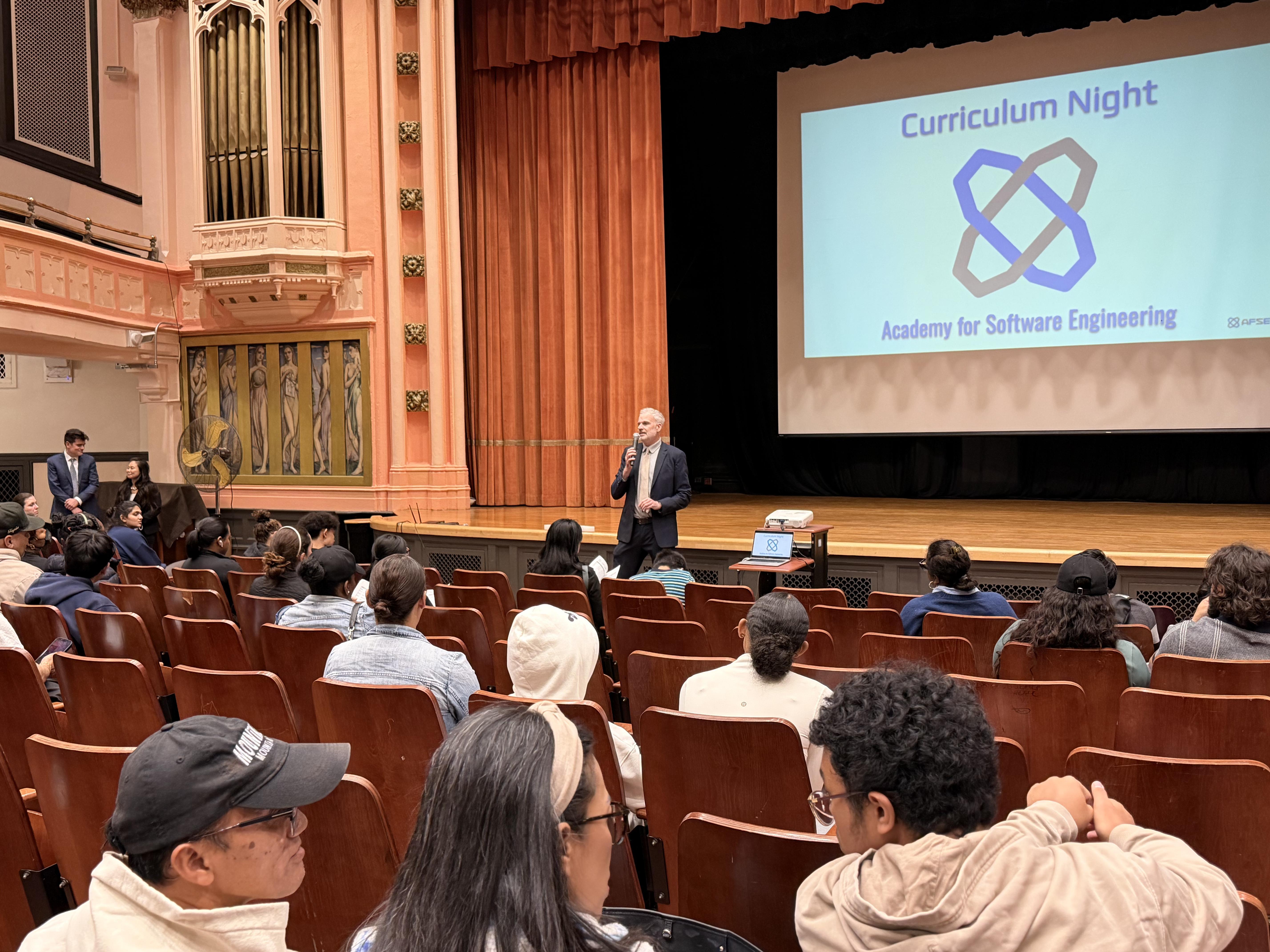 A speaker addresses a seated audience at a Curriculum Night event in an auditorium.