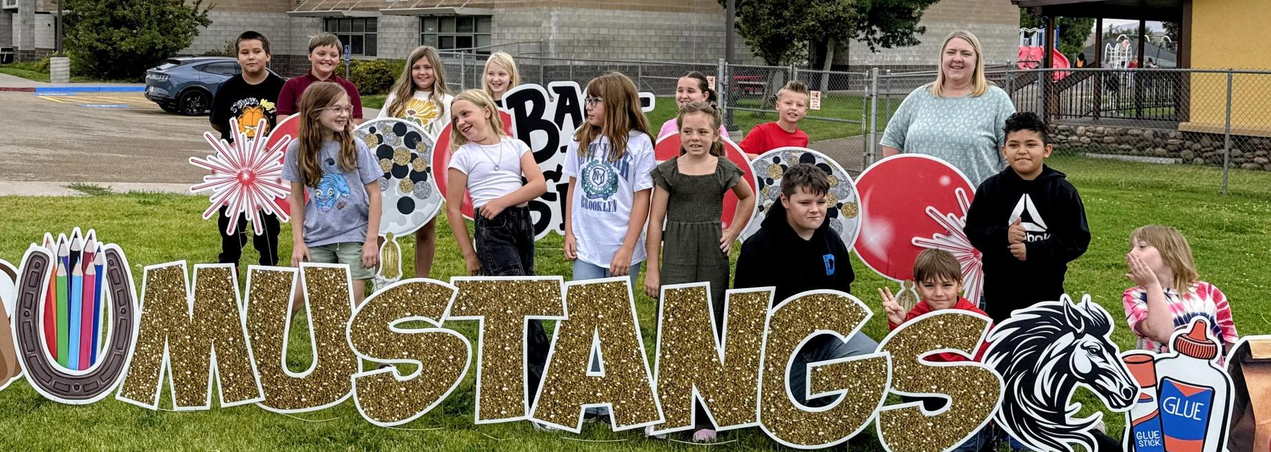 Group of children posing around a large 'Mustangs' sign outside a building.