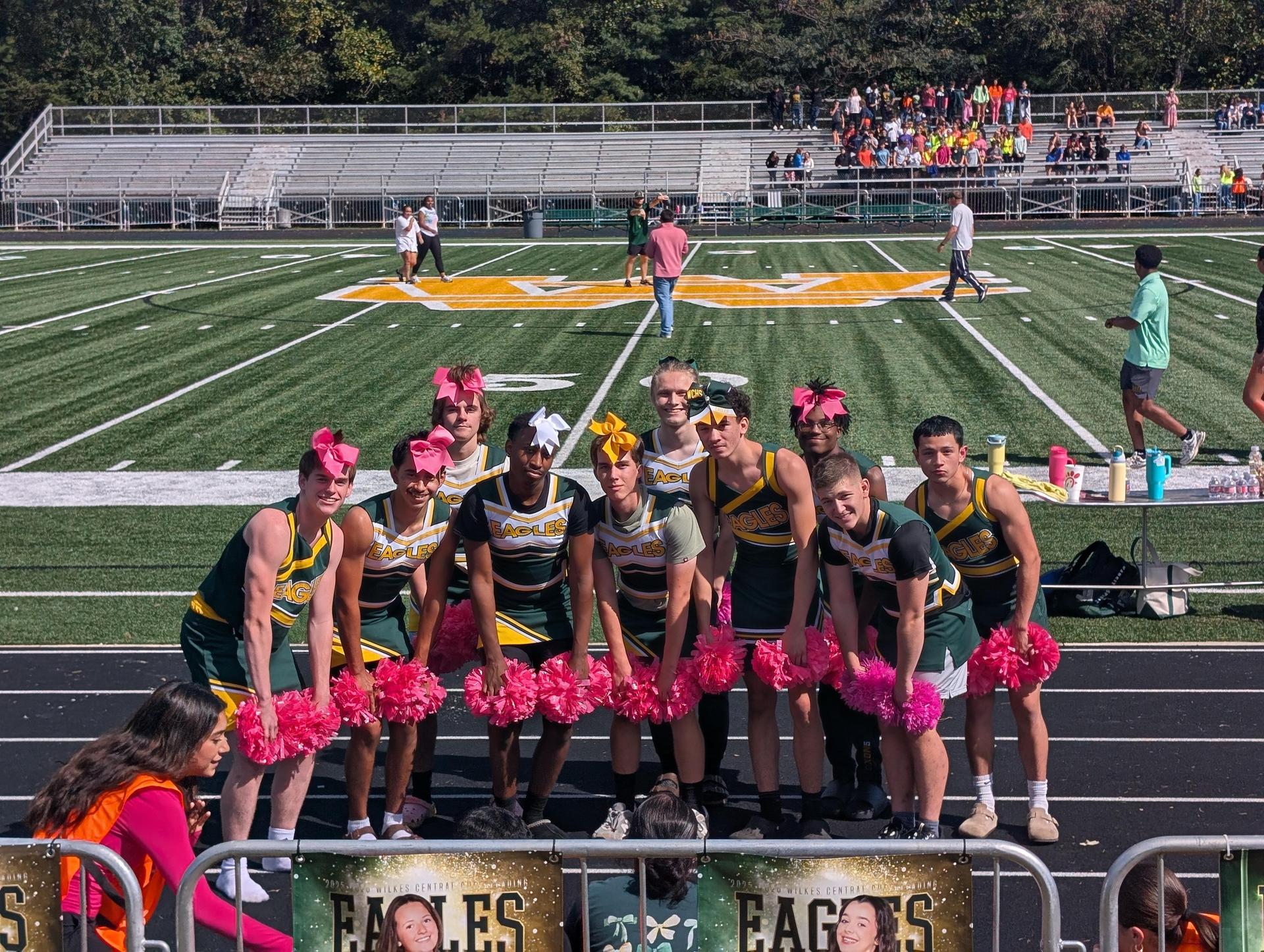 Cheerleaders in colorful outfits pose with pink pom-poms on a sports field.