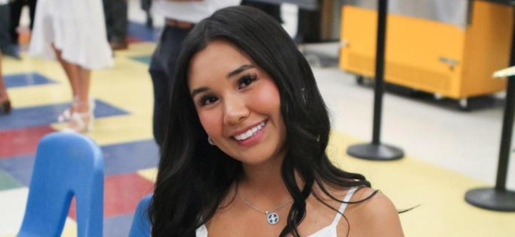 Graduate in white dress holding blue roses, seated in a colorful area.