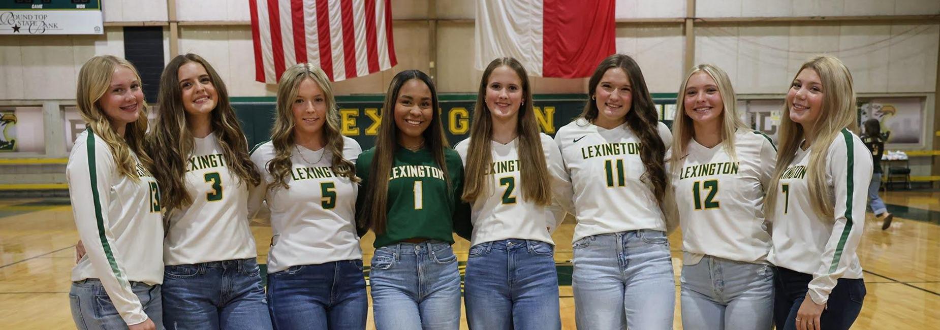 Group of female athletes in team jerseys and jeans, standing on a gym floor.