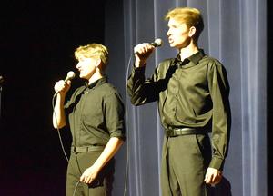 Two young men in black shirts sing into microphones on stage.