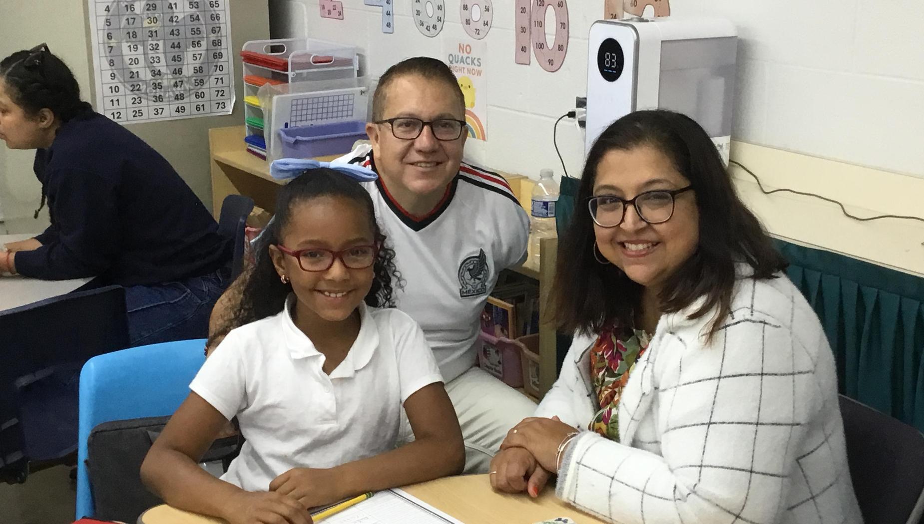 A smiling girl with glasses and two adults at a classroom table.