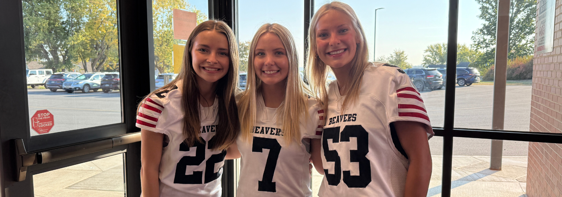 Three girls in football jerseys smiling at a glass door.