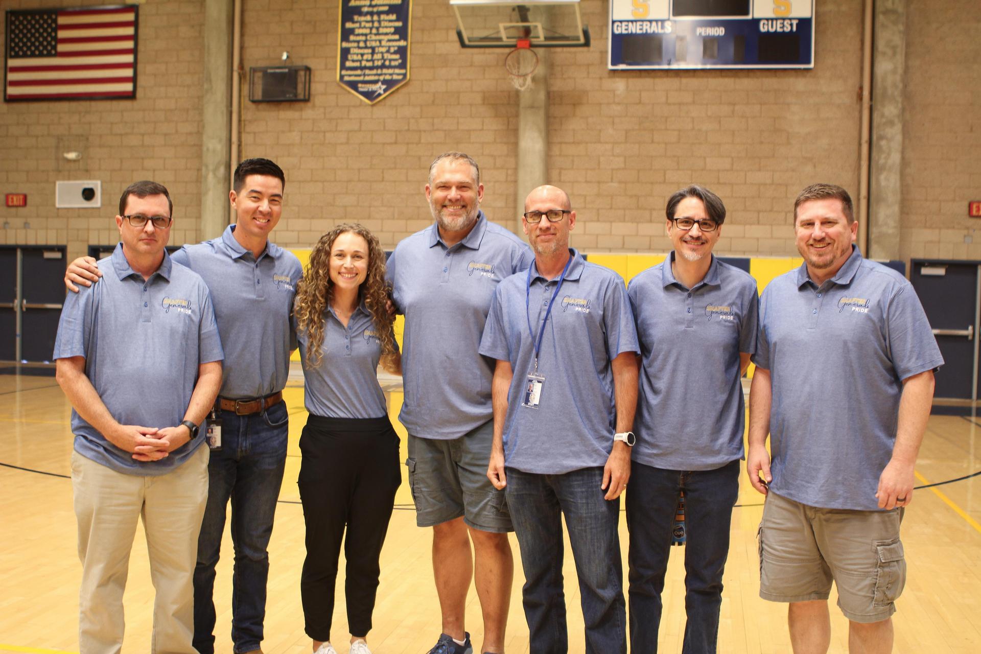 A group of seven people standing together in a gymnasium, all wearing matching blue shirts.