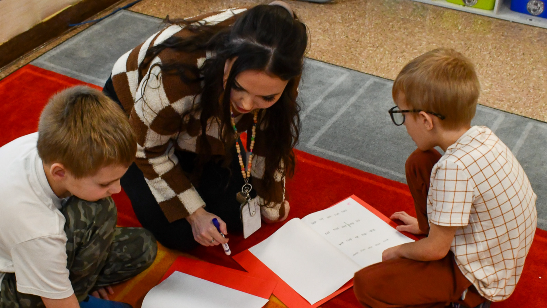 Teacher assisting two children with worksheets on a classroom carpet.