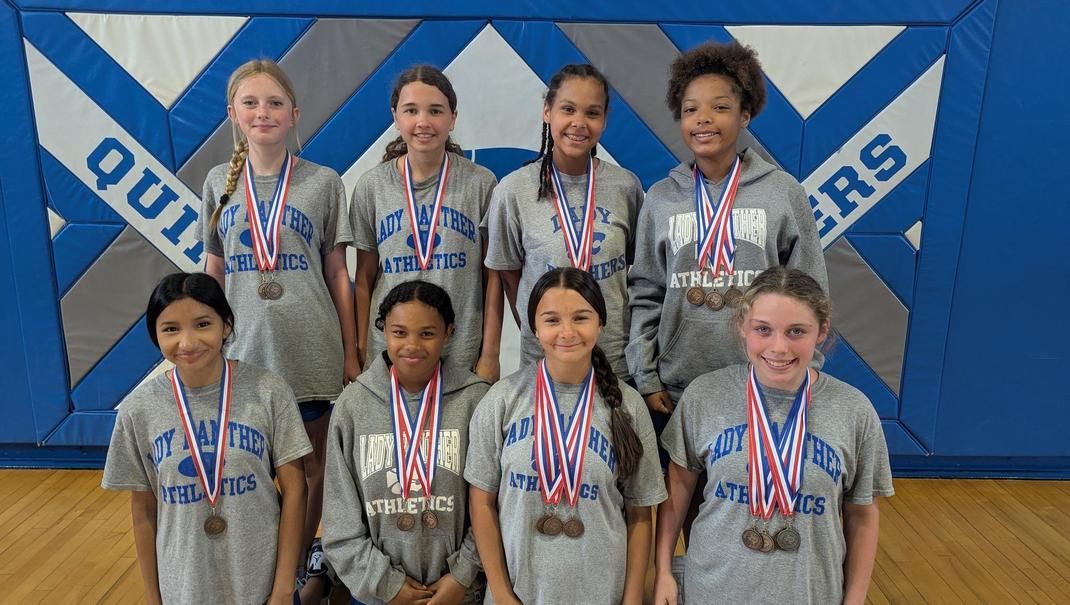 Group of eight young athletes wearing gray shirts with medals on a gym floor.