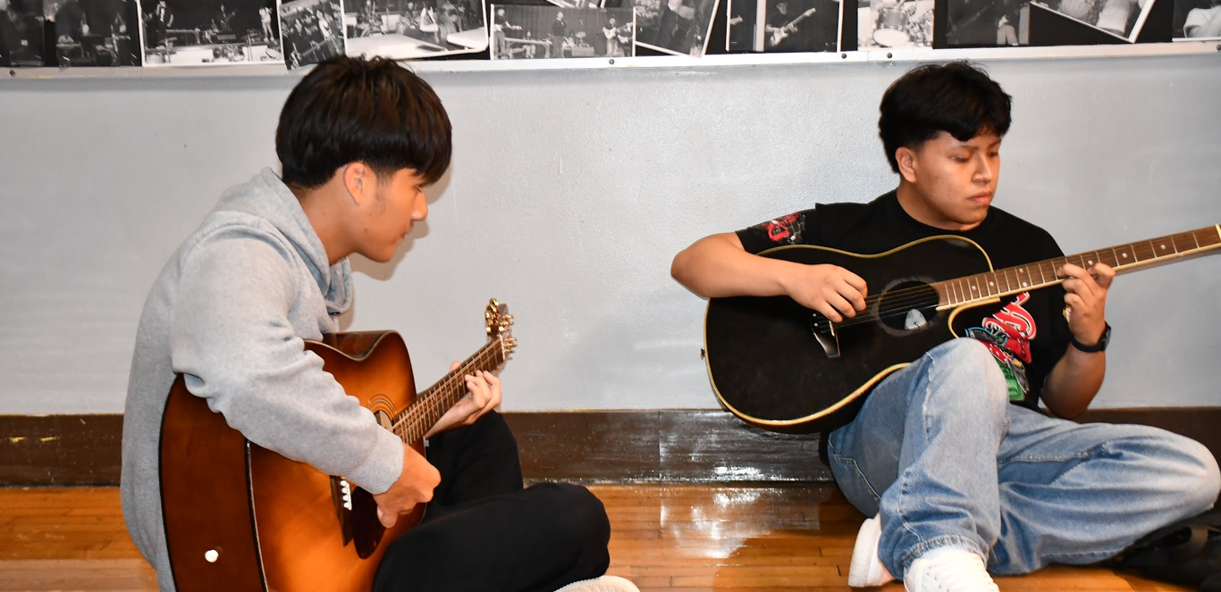 Two students practicing playing guitar on the floor in the hallway.