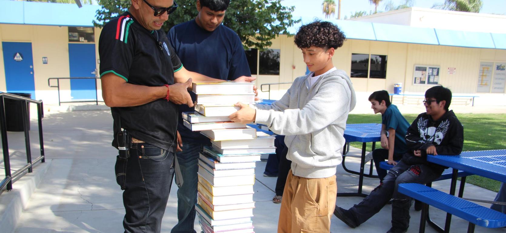 A student stacking books with assistance outdoors near picnic tables.