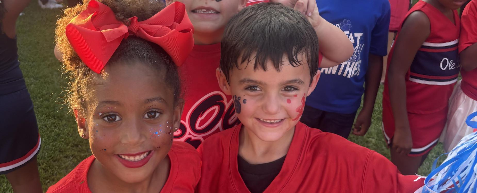 Three young children smiling at the camera, wearing matching red shirts and face paint.
