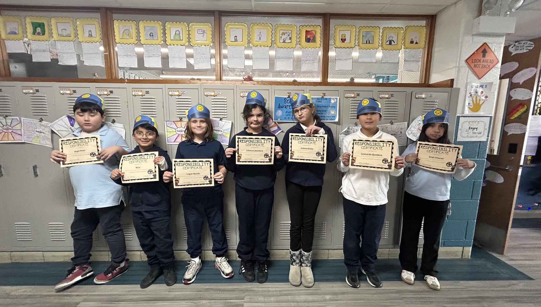 Seven students wearing hats and holding certificates in front of lockers.