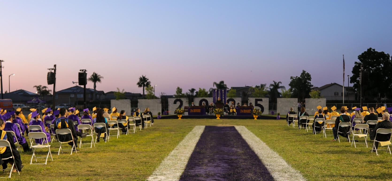 Graduation ceremony with graduates in purple and gold gowns, seated and waiting.
