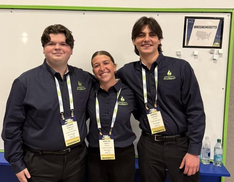 Three Taunton High School students wearing matching Massachusetts ProStart shirts and competition badges stand together smiling in front of a whiteboard.