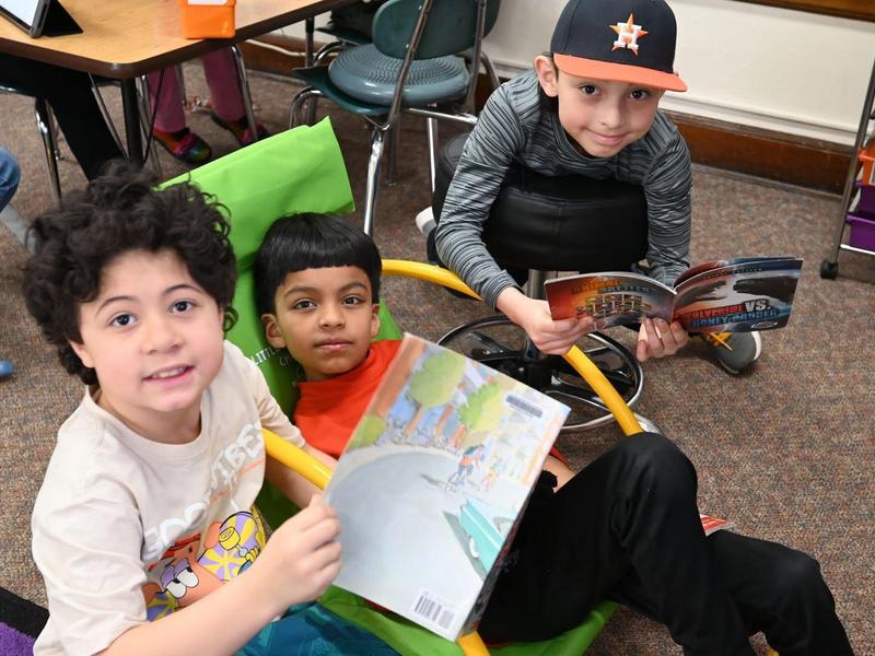 Three children in a classroom, reading books and sitting in colorful chairs.