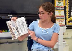 Girl reading from a book to a class while sitting in front of a screen.