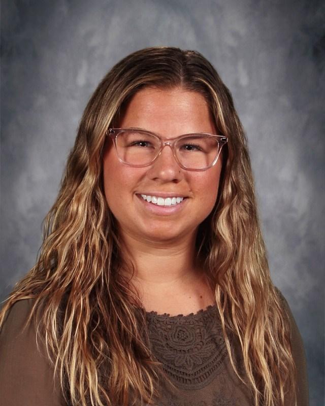 Smiling woman with wavy brown hair and glasses in a patterned top.