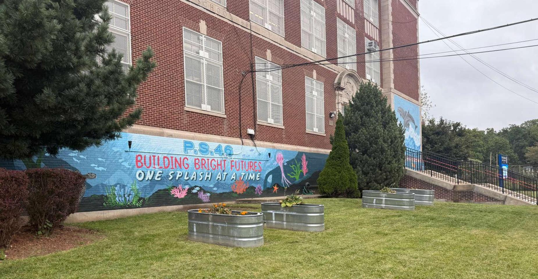 Colorful mural on a school wall with painted flowers and text, metal planters in a grassy area.