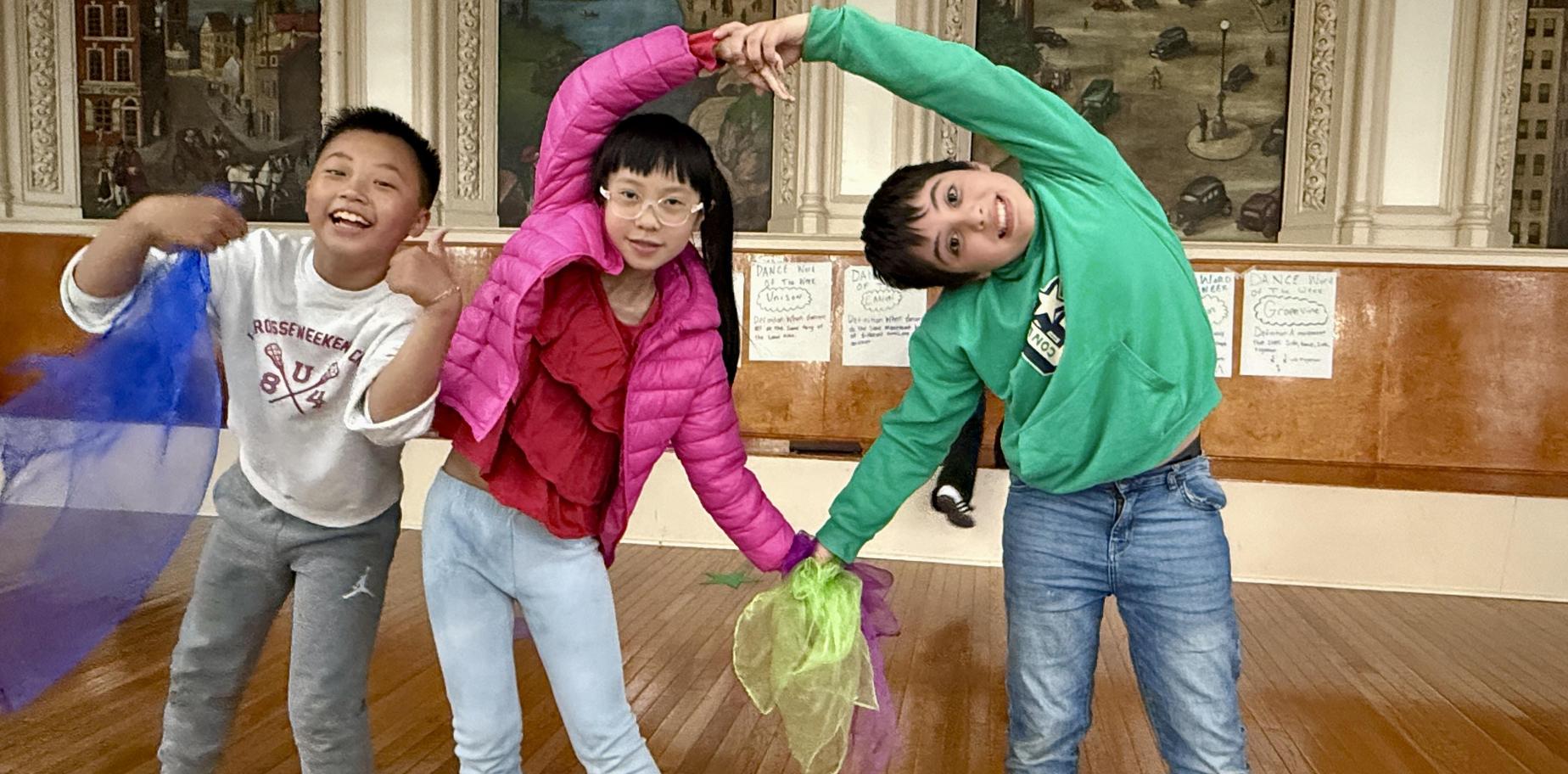 Three children playfully posing with colorful scarves in a dance studio.