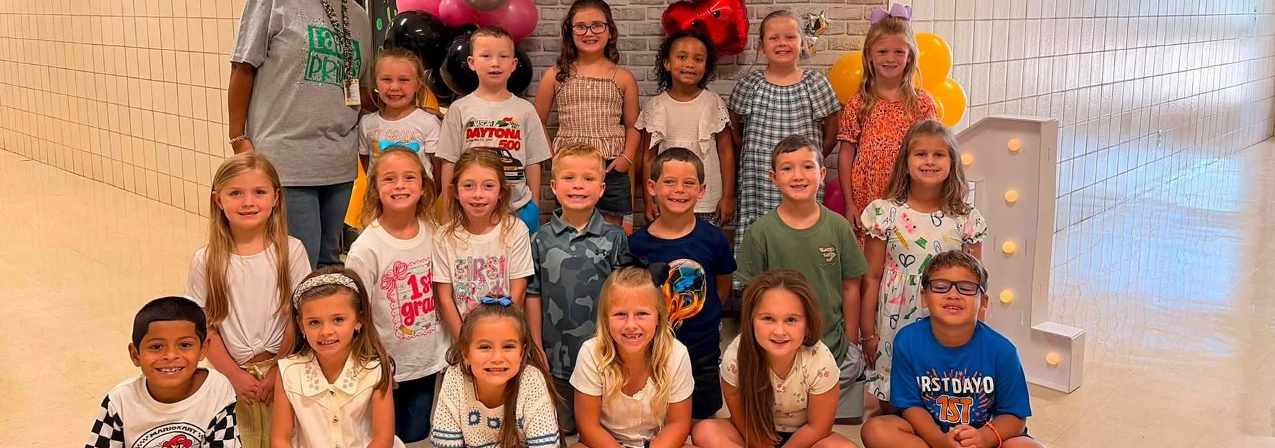 Group of children and an adult in front of colorful balloons and a wall decoration.