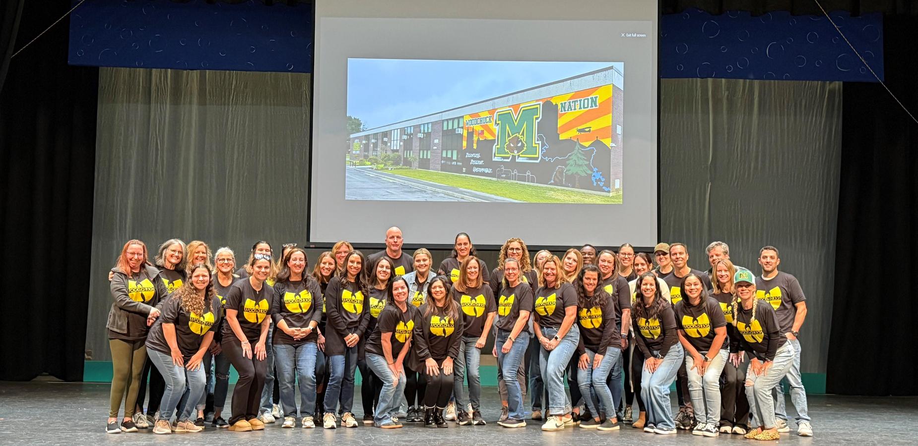 A large group of Martin Middle School staff members pose together on stage during a professional development day. They are all smiling and wearing matching black shirts with a yellow “Woodchuck Nation” logo. Behind them, a projector screen displays an image of the school building with a colorful mural featuring the Martin Middle School “M” and the words “Woodchuck Nation.” The group stands in front of a blue and gray stage backdrop, reflecting a sense of unity and school pride.