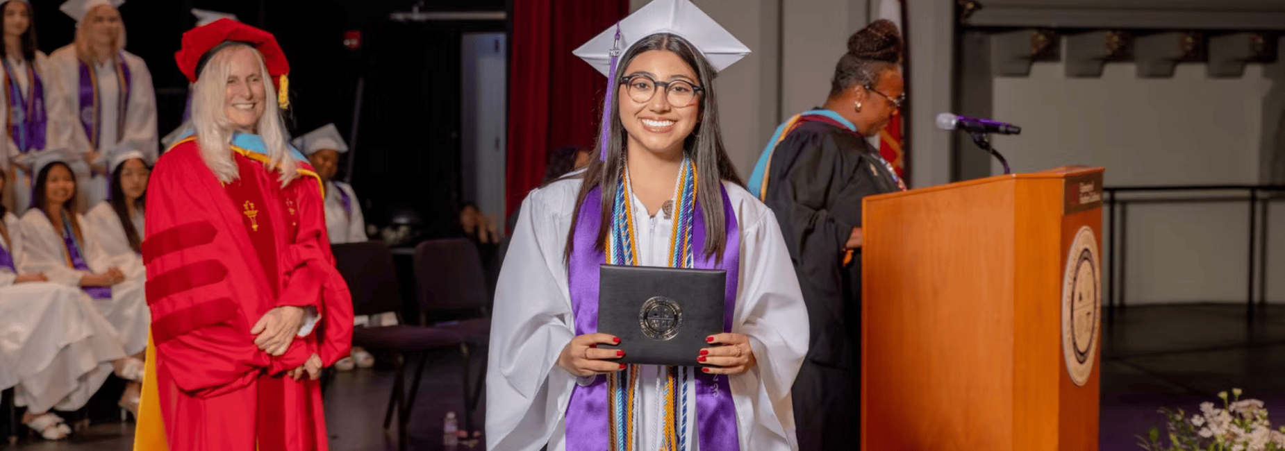 Graduate in cap and gown holding diploma on stage with faculty and classmates behind.