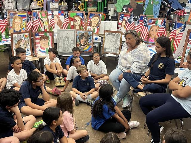 A teacher speaks to a diverse group of students sitting on the floor in an art classroom.