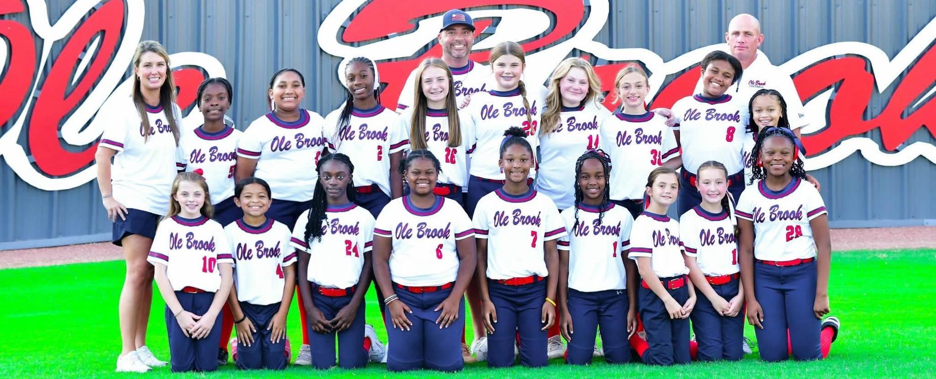 A youth softball team poses in matching uniforms in front of a large logo.