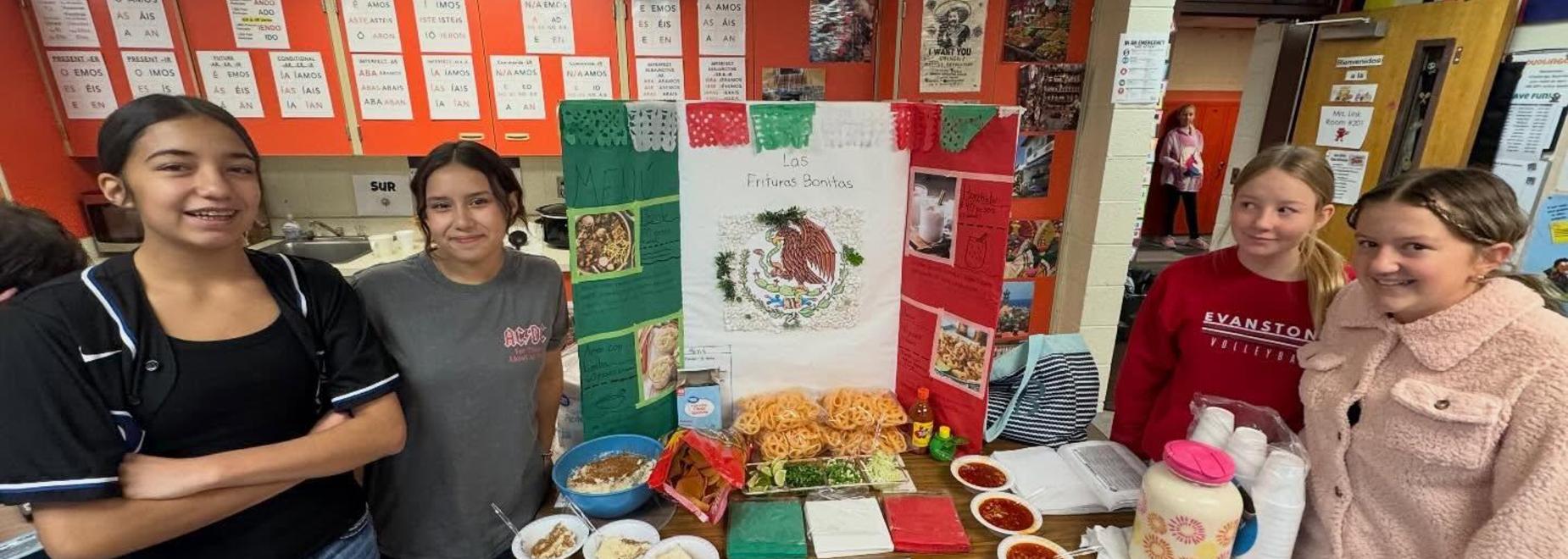 Four girls stand in front of a colorful poster featuring food and cultural decorations.
