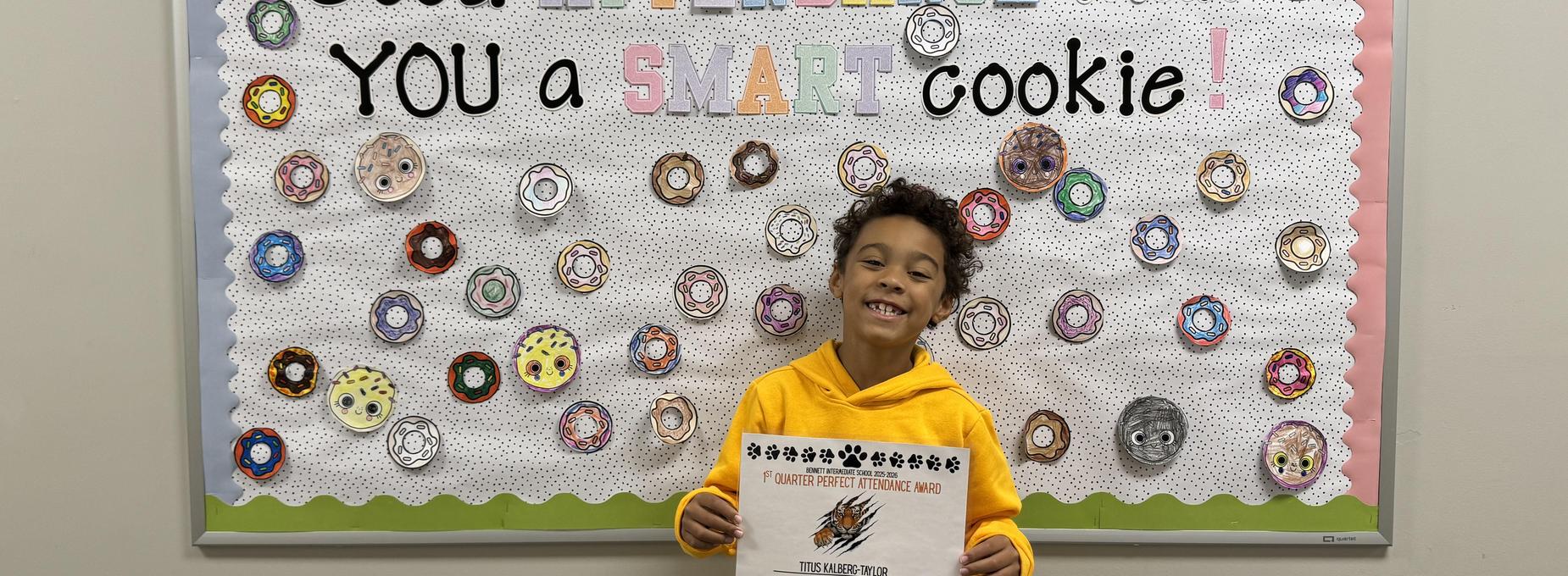 A smiling child in a yellow hoodie holding a certificate in front of a colorful wall display.