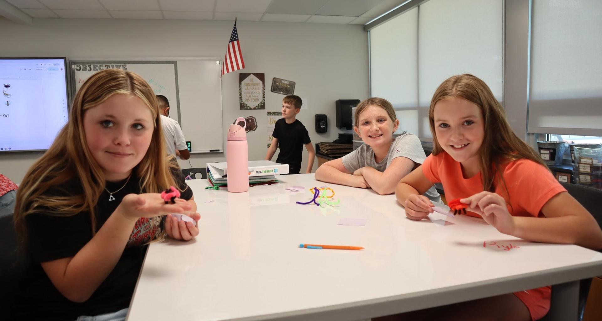 Three girls smile at the camera while working on creative projects at a classroom table.