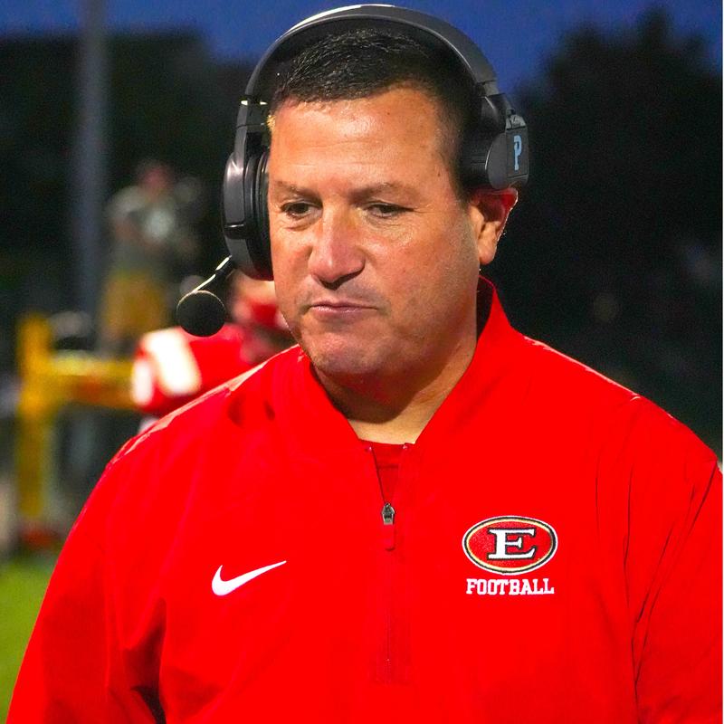 Coach wearing a red jacket and headset, looking contemplatively on the field.