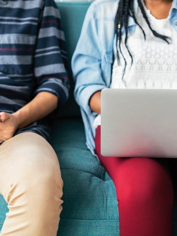 Two people sitting on a couch, one using a laptop, showing their lower bodies.