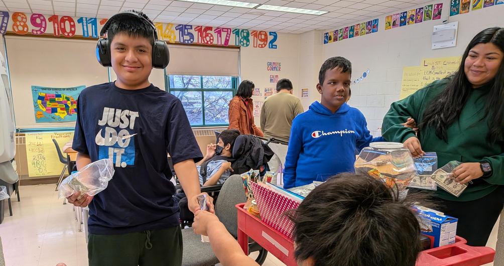 Students interacting in a classroom during a hands-on activity, with one boy passing money to another.