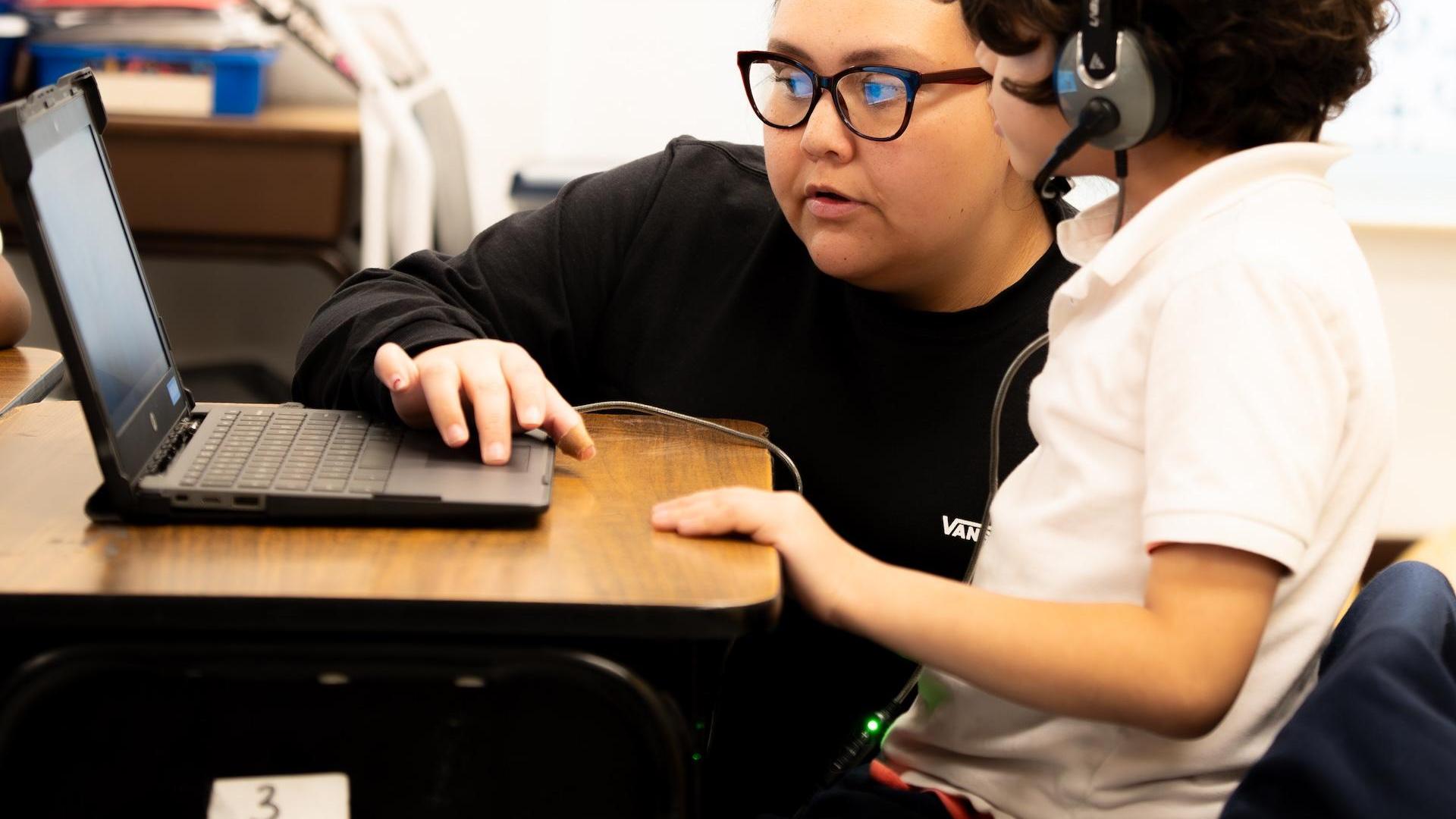 A child and an adult focused on a laptop while wearing headphones.