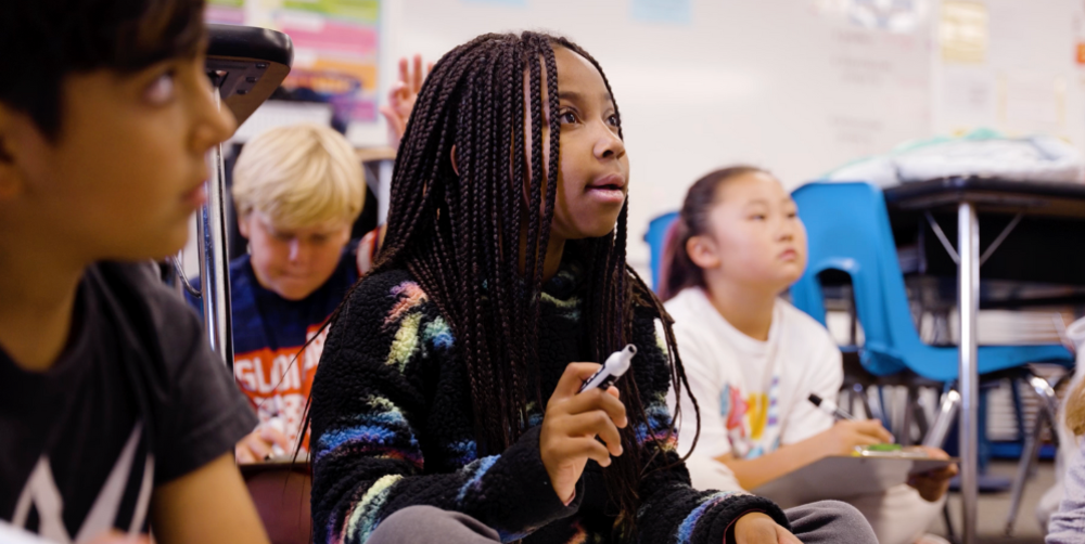 Focused students in a classroom, with one girl raising her hand to speak.