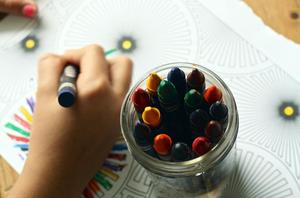 A child's hand holding a crayon, coloring next to a glass jar of crayons.