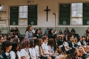 Students in a hall attending a ceremony with a cross on the wall.