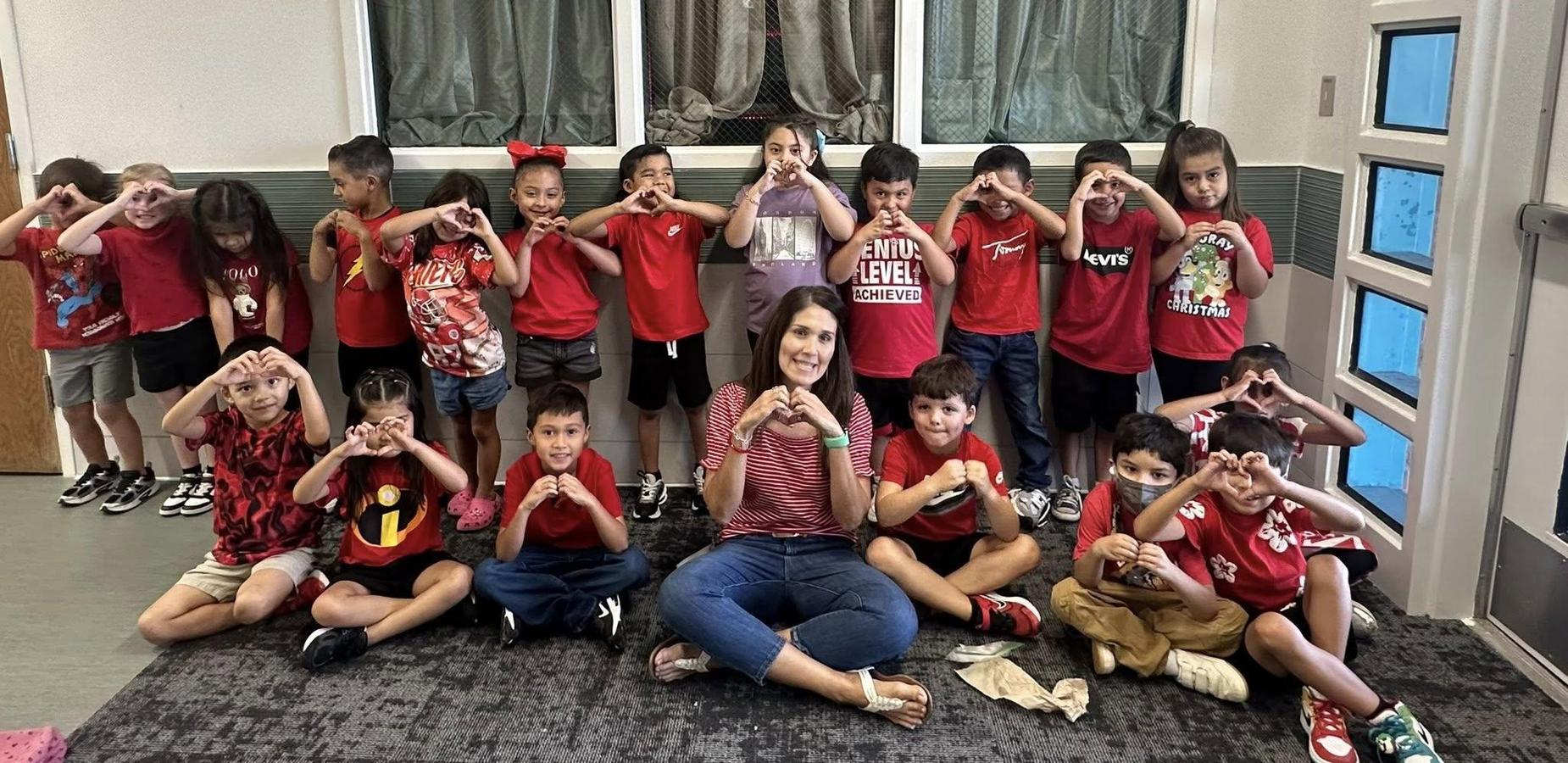 class of students in red shirts standing and sitting around teacher; all making hearts with hands