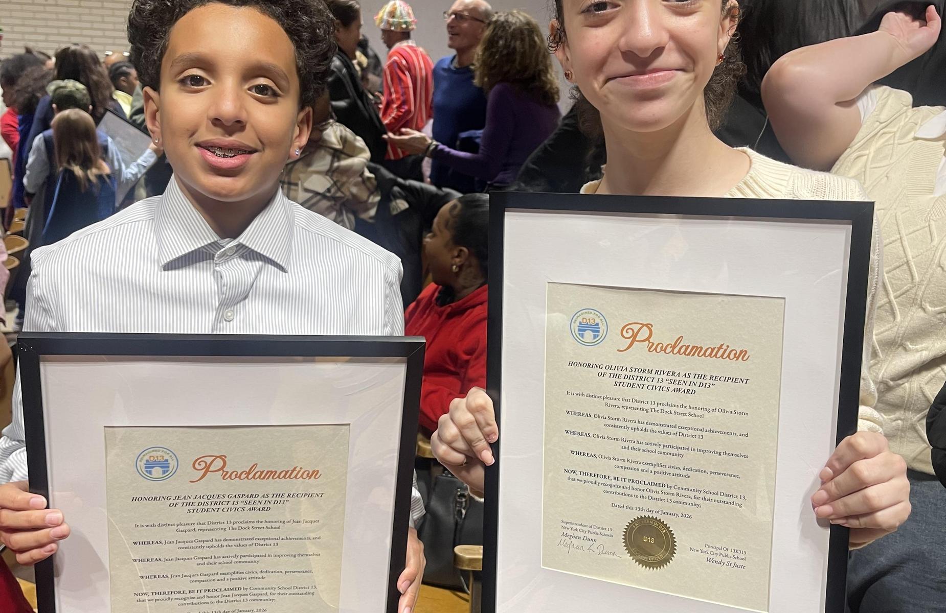 Two children holding framed proclamations with smiles in a crowded indoor setting.