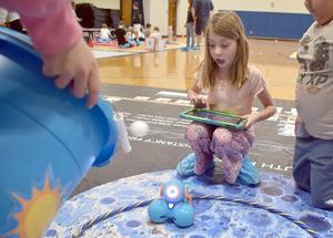 A girl uses a tablet to control a robot on a colorful mat in a gym.
