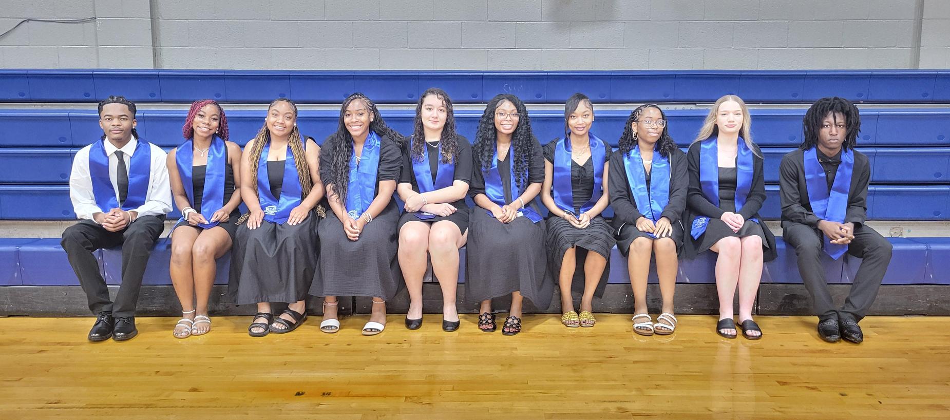 Row of students in blue and black attire seated on the bleachers.