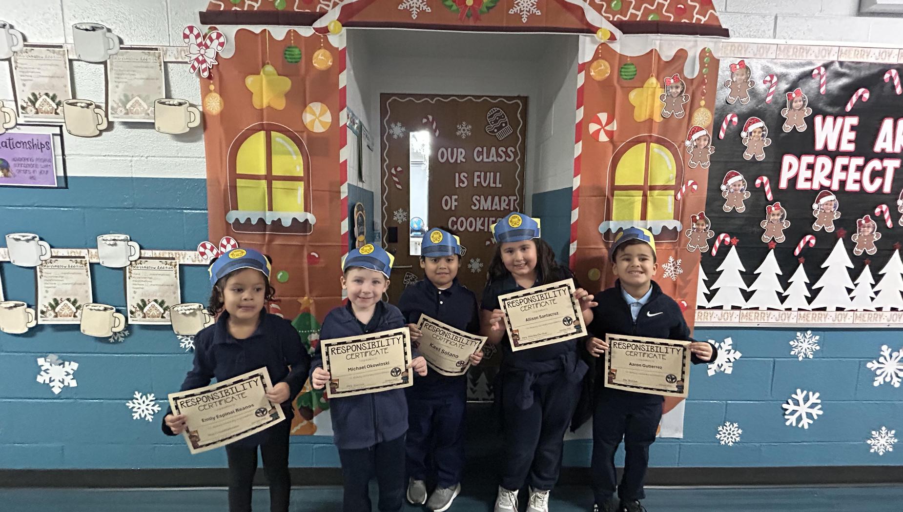 Five children in blue outfits holding certificates in front of a decorated gingerbread house.