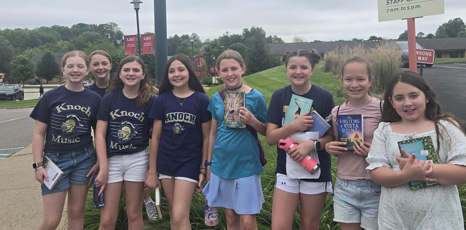Group of girls holding books, smiling outside a school parking area on a cloudy day.