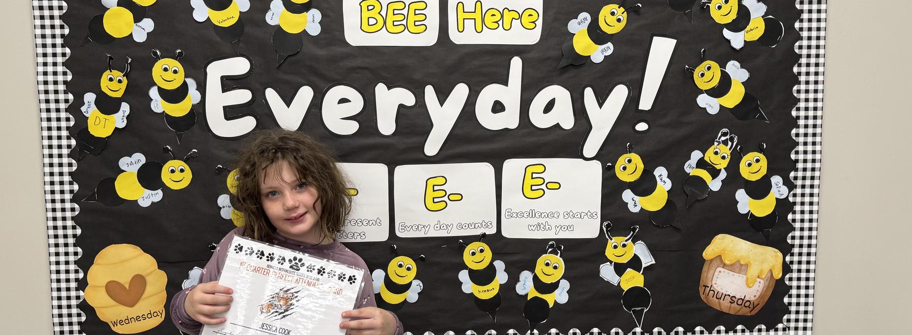 Child holding a certificate in front of a bee-themed bulletin board.