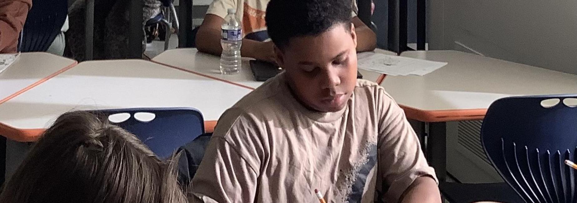 A student focused on drawing at a classroom desk, with other students in the background.