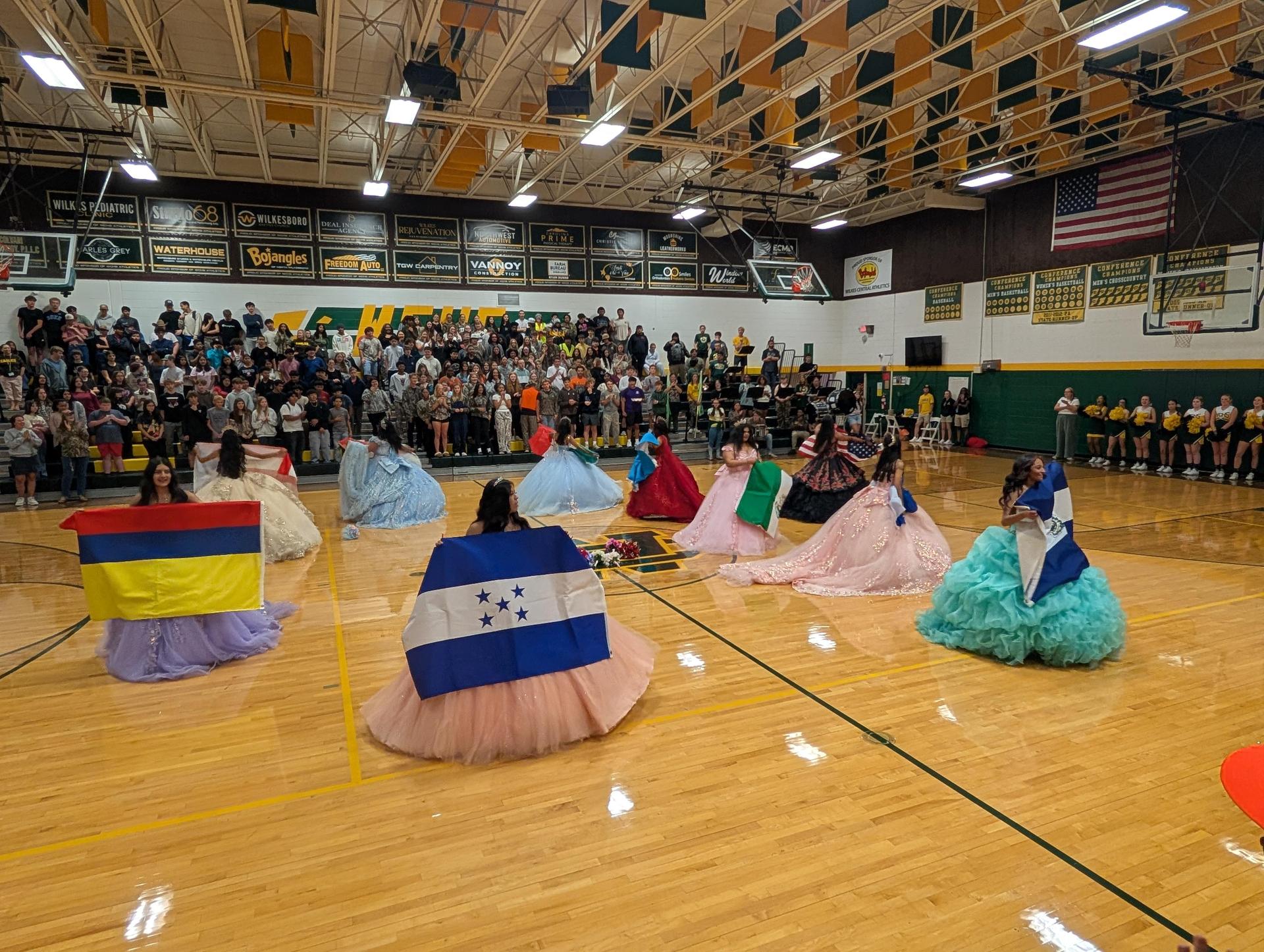 Girls in vibrant ball gowns with flags representing different countries, dancing in a gymnasium.