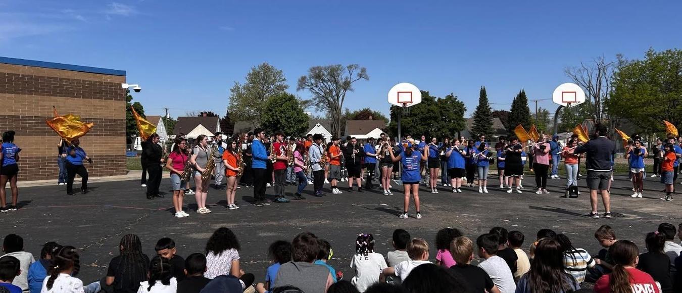 Students sit on the asphalt watching a school marching band perform on an outdoor basketball court.