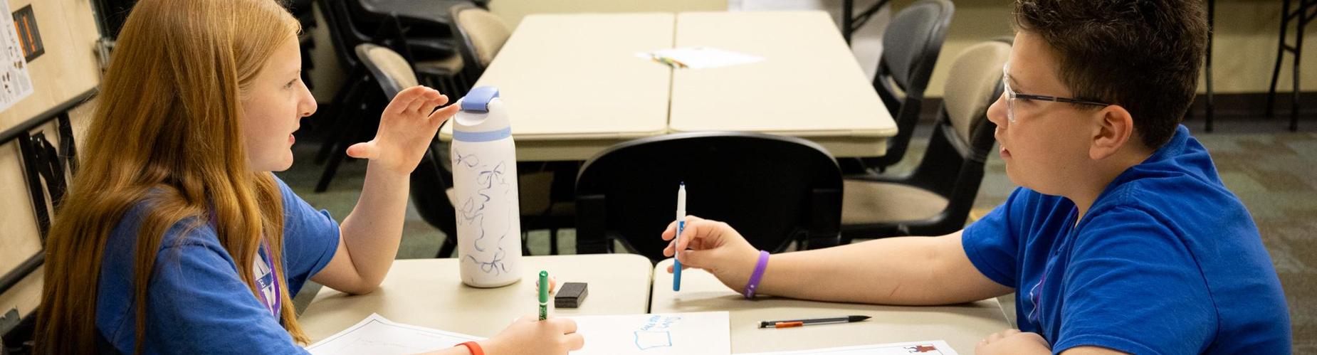 Two children in blue shirts sit at a table engaged in conversation with drawing materials.