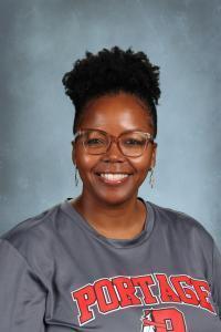 Woman with curly black hair in glasses wearing a gray shirt with text smiling against a gray background.