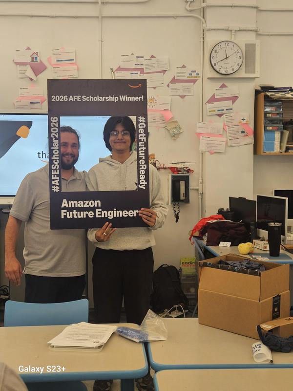 Two people posing with an award sign in a classroom filled with materials.